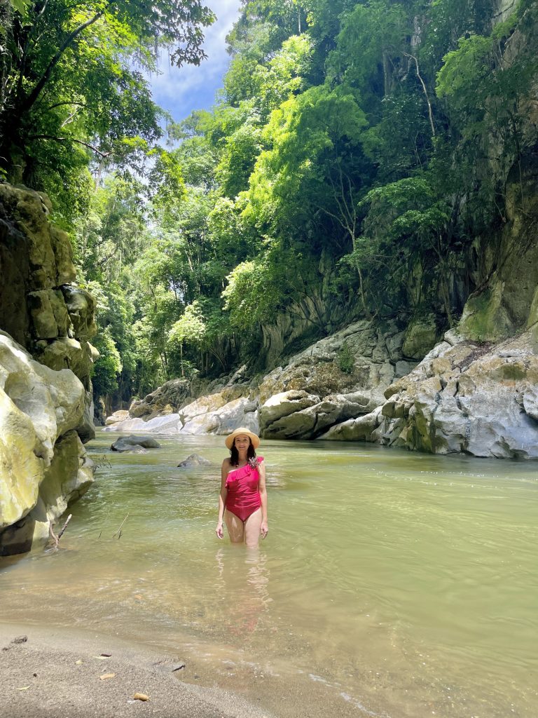 Traveler wading in a calm shallow area of El Boquerón Guatemala canyon with limestone rocks, green river water, and jungle around her.