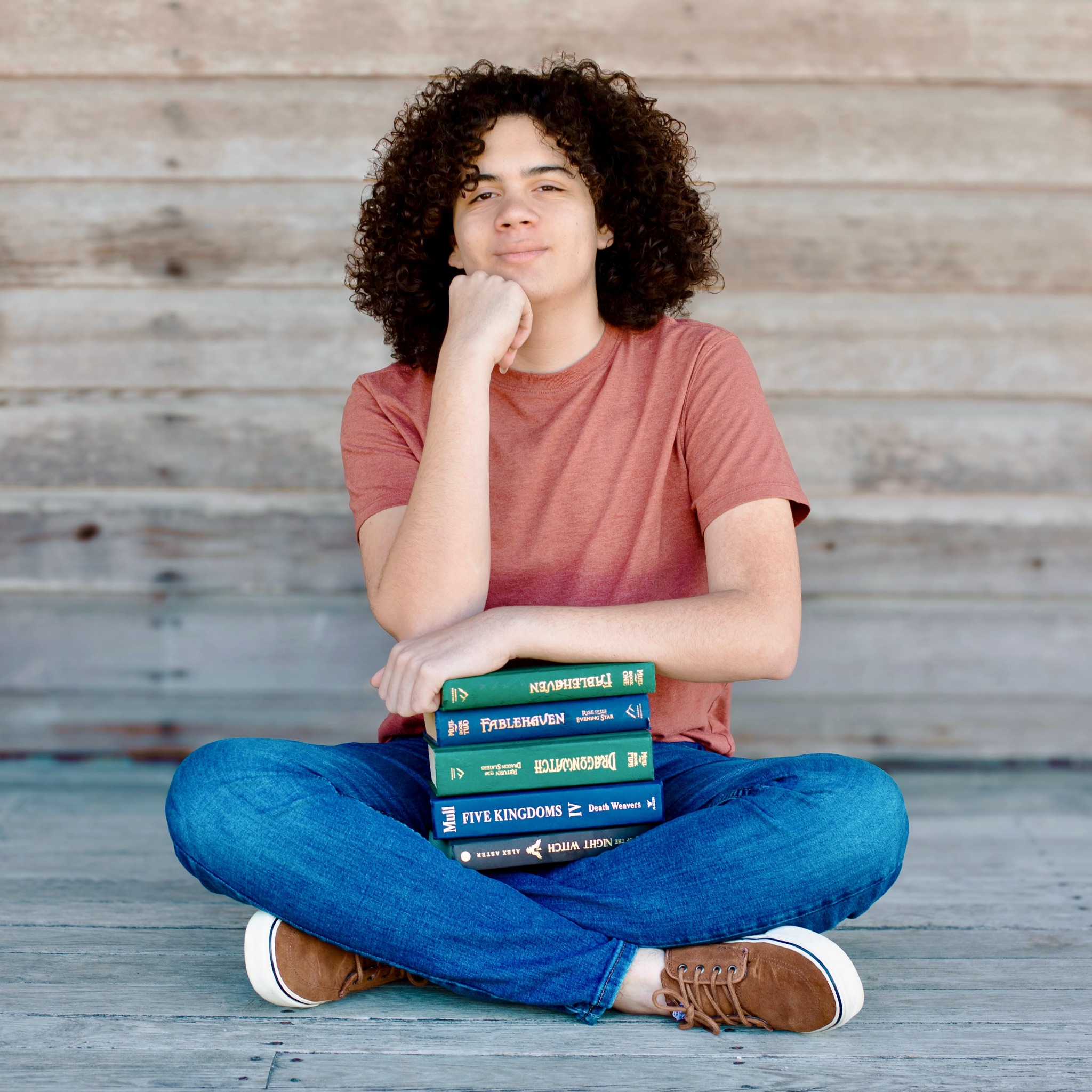 Teenage boy sitting cross-legged with a stack of fantasy novels including Fablehaven and Dragonwatch
