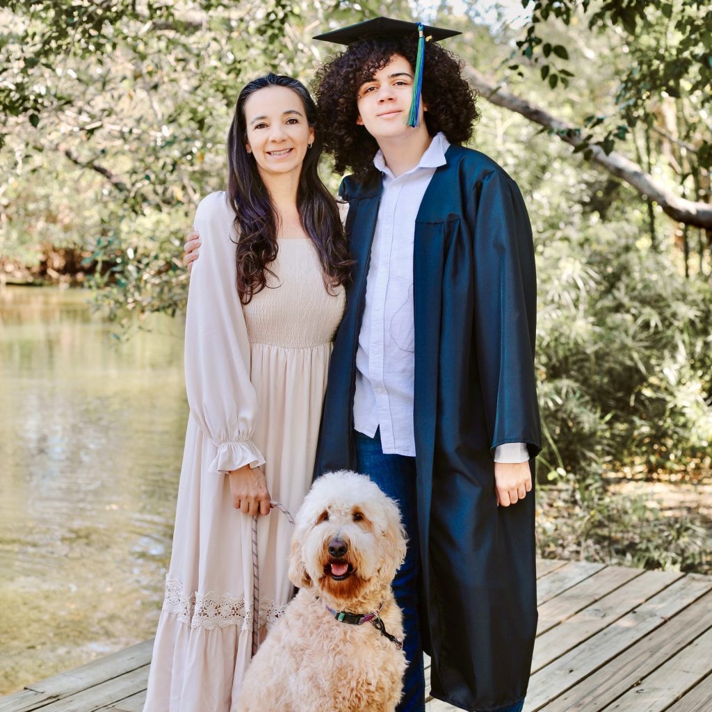 Mother and son smiling with their goldendoodle on a dock, son wearing a graduation cap and gown