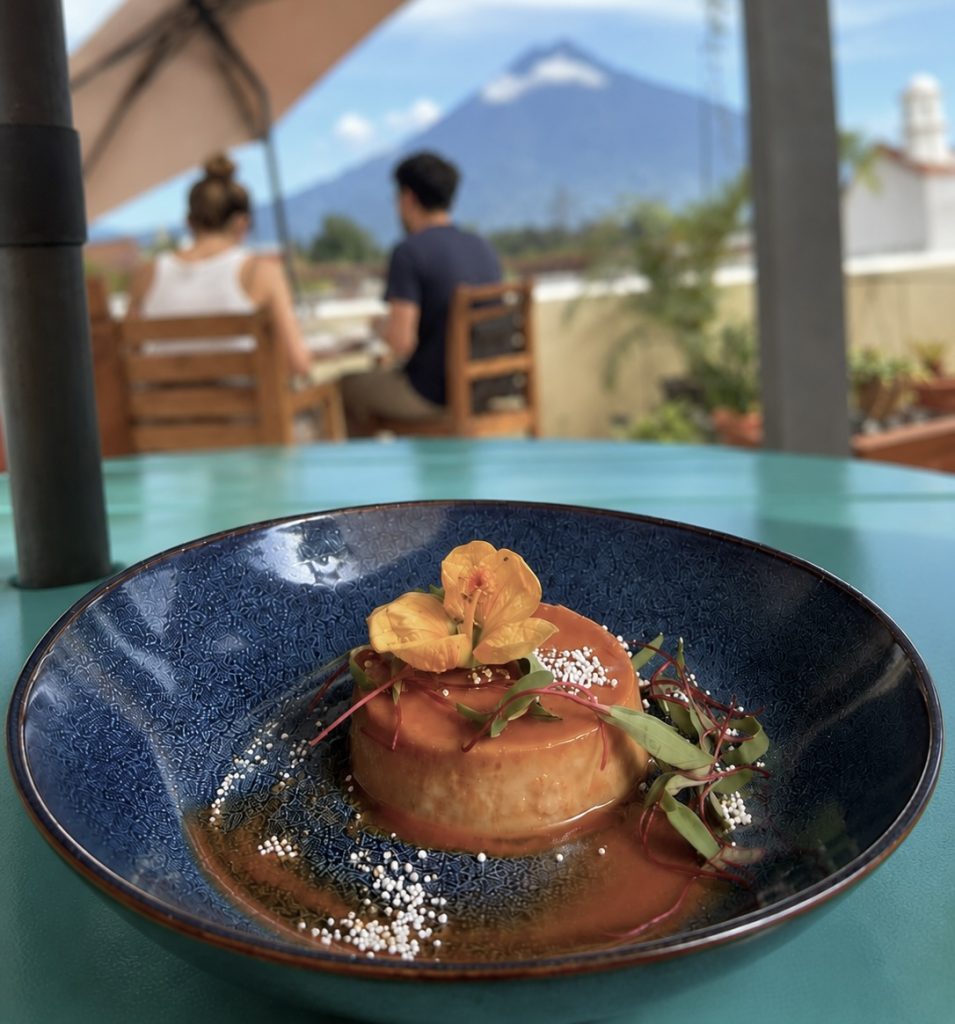 Caramel flan topped with edible flowers at 27 Adentro in Antigua Guatemala, with Volcán de Agua in the background.